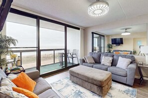 A spacious living room with a large window, a grey sofa, an ottoman, and a flat-screen TV. - Adorable beachfront condo with balcony, incredible views, pools & game room (Ocean City)