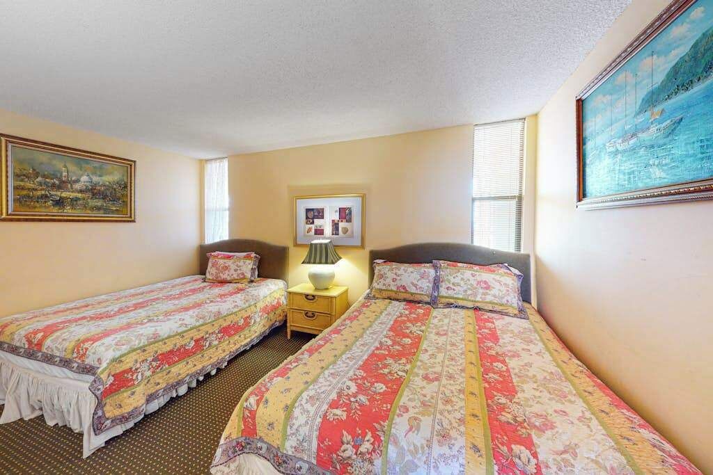 Une chambre d’hôtel avec deux lits simples, une table de chevet avec une lampe, un tableau encadré au mur et une fenêtre avec des stores.