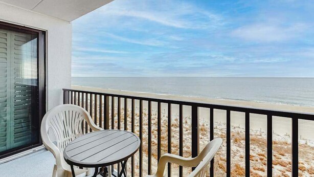 Ein Balkon mit Tisch und Stühlen, der auf einen Strand mit freiem Blick auf das Meer blickt.