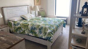 Ein Schlafzimmer mit Bett, Nachttisch, Lampe, Fenster mit Rollläden, einem gerahmten Bild an der Wand und einem Regal mit Dekorationen.