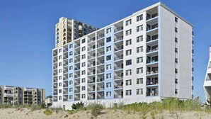 Ein mehrstöckiges Hotel mit Balkonen, einer flachen Dachterrasse und einem Sandstrand im Vordergrund.