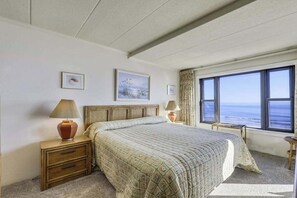 A bedroom with a bed, two bedside tables, a window with a view of the sea, and a painting on the wall. - Attractive Suite in Oceanfront Complex w/Free WiFi, Private Washer/Dryer, & AC (Ocean City)