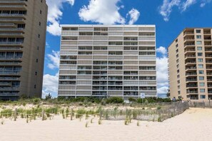 A modern hotel with a grid of balconies, a sandy beach in the foreground, and other buildings in the background. - Beachfront condo with indoor pool, new fitness center, ocean & beach views (Ocean City)