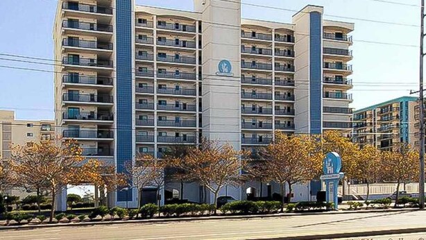 A multi-story hotel with a central tower, balconies, and a parking area with trees. - Excalibur 303 (Ocean City)