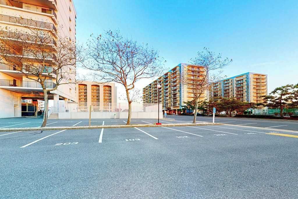 Un estacionamiento con espacios delimitados, un edificio de varios pisos y un árbol en primer plano.