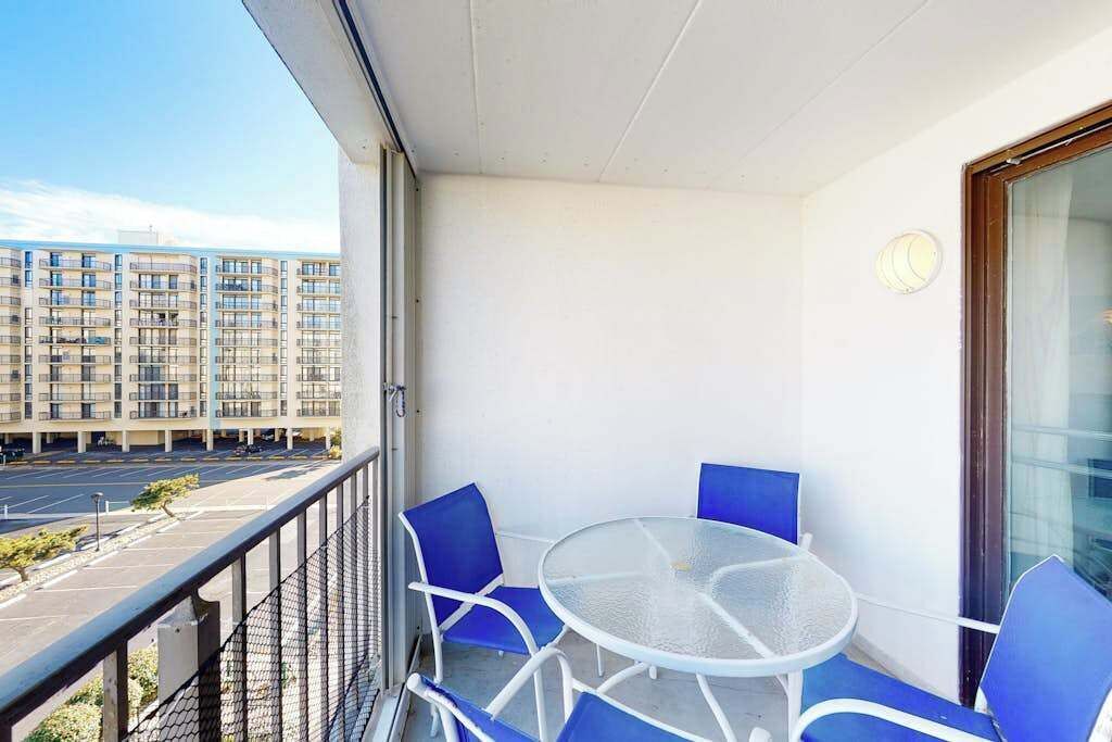 A balcony with a glass table and blue chairs, overlooking a parking lot and a multi-story building.