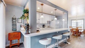 A modern kitchen with white cabinets, a central island with bar stools, pendant lighting, and a dining area with wooden chairs. - Drawbridge Rd 38 (Ocean Pines)