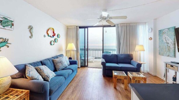 A living room with a blue sofa, a blue armchair, a ceiling fan, and a view through a glass door to the outside. - Beautiful Home in Oceanfront Building w/Shared Pool, Free WiFi, Central AC (Ocean City)