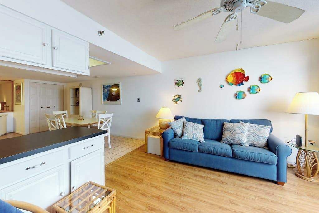 A modern living room with a blue sofa, wooden flooring, white cabinets, and a ceiling fan.
