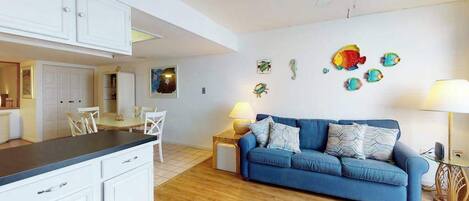 A modern living room with a blue sofa, wooden flooring, white cabinets, and a ceiling fan.