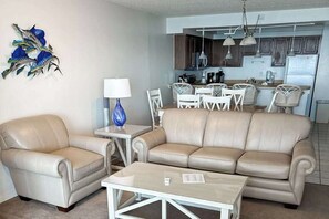 Ein Wohnzimmer mit einem beigen Sofa, einem Couchtisch und einem Essbereich mit weißen Stühlen.