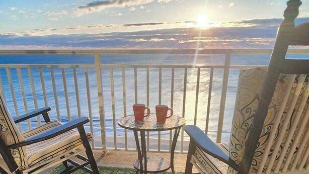 Ein Balkon mit Blick auf das Meer, zwei Stühle, ein kleiner Tisch mit zwei roten Tassen und ein stuhl mit Muster.