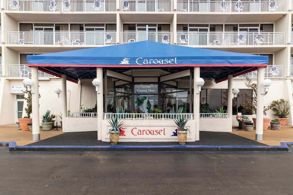 Entrada de hotel con toldo azul identificado como 'Carousel', plantas en macetas y un balcón con barandillas blancas.