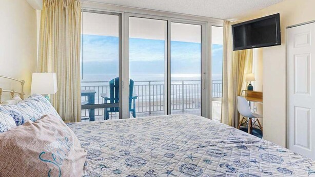 A hotel room with a bed, a desk, a chair, a TV, and a balcony with a view of the sea. - Carousel 807 (Ocean City)