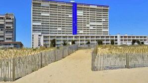 A large hotel with multiple floors, a sandy pathway leading to the entrance, and a wooden fence on either side. - Carousel 807 (Ocean City)