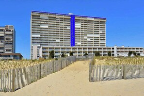 A large hotel with multiple floors, a sandy pathway leading to the entrance, and a wooden fence on either side. - Carousel 807 (Ocean City)