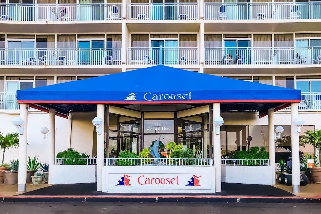 Entrada de hotel con un toldo azul con la leyenda 'Carousel', un letrero que indica '11700 Coastal Hwy' y un balcón con barandas blancas.