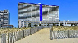 Een groot hotel met meerdere verdiepingen, een opvallende blauwe verticale streep en een zandpad dat naar de ingang leidt.