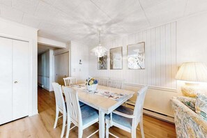 A dining area with a wooden table, white chairs, a chandelier, and wall art. - Top-floor, oceanfront condo w/ gorgeous view, balcony, beach access, & WiFi (Ocean City)