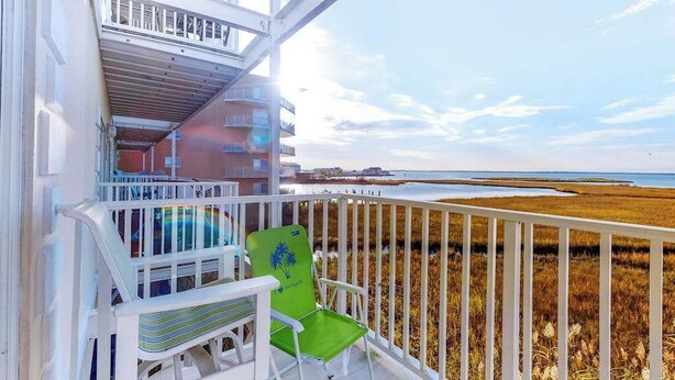 A balcony with a view of a beach and marshland, a green chair, and a striped chair. - Roomy & convenient bayfront home with pool, central AC & balcony (Ocean City)