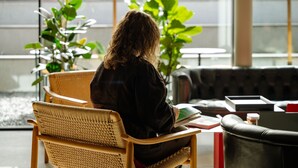Une personne est assise sur une chaise en osier dans une pièce d’où l’on aperçoit une rue et des plantes à l’extérieur.