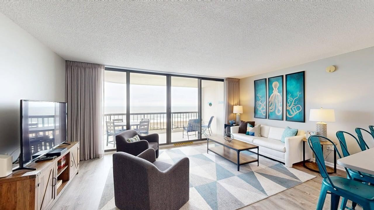 A hotel room with a balcony, a TV, a sofa, a dining table, and artwork on the wall.