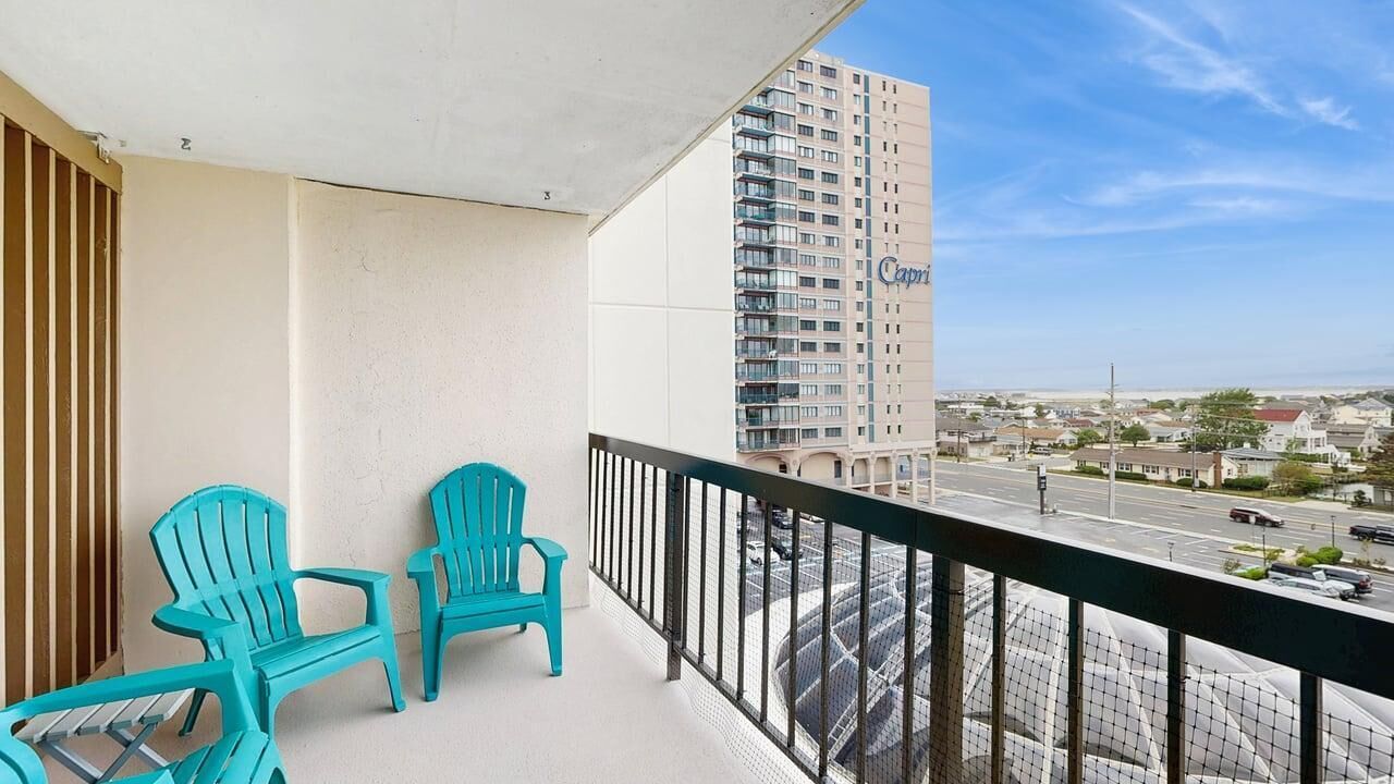 Un balcon avec deux chaises turquoise, une vue sur un grand immeuble portant l’inscription « Capri », et une rue avec des voitures.