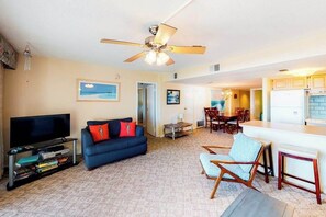 A living room with a television, sofa, and chairs. There is a ceiling fan, a dining area, and a kitchen visible in the background. - Irene 404 (Ocean City)