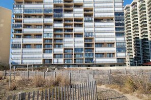 A multi-story building with a grid-like facade, balconies, and a sandy ground area with a wooden fence. - Oceanfront condo with balcony, AC, pool, & W/D (Ocean City)