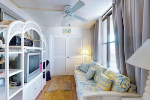A room with a white sofa, a ceiling fan, a TV on a white entertainment unit, and a door with shutters. - Oceanfront condo with balcony, AC, pool, & W/D (Ocean City)