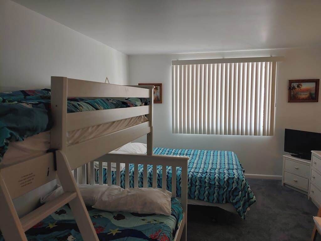A room with bunk beds, a window with blinds, a television, and a dresser.