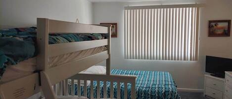 A room with bunk beds, a window with blinds, a television, and a dresser.