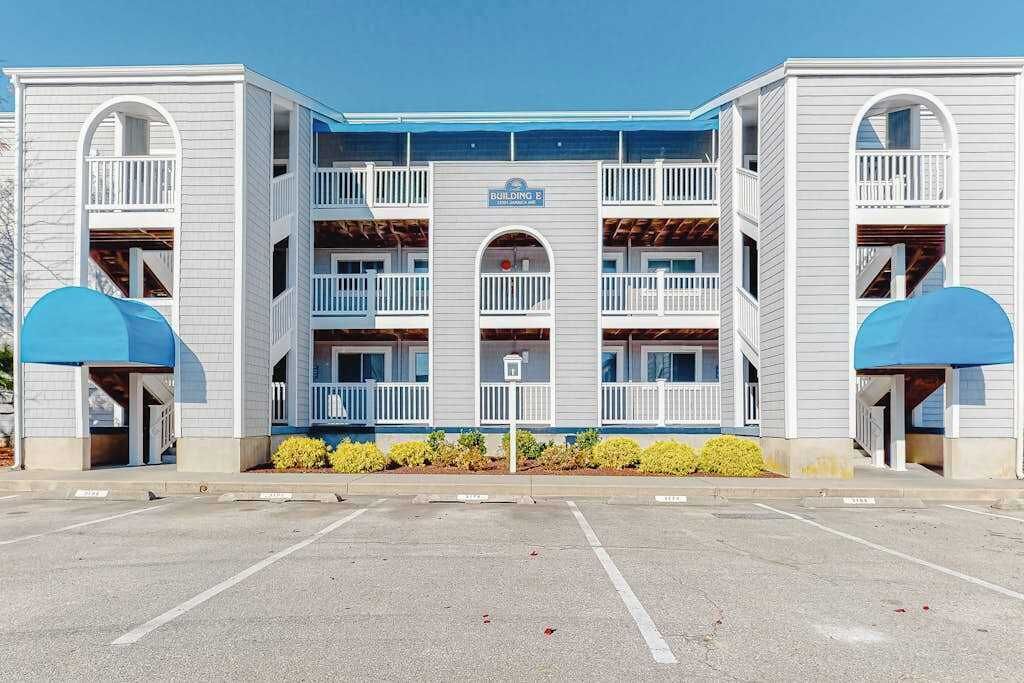 A multi-story building with a central entrance, balconies, and a parking lot in front.