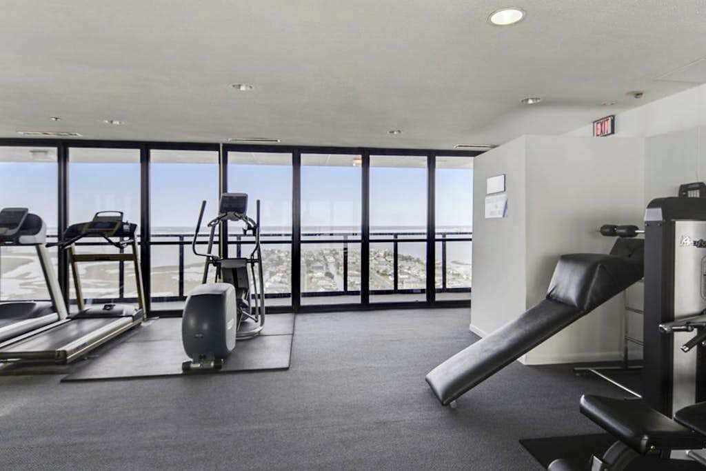 Une salle de sport équipée de tapis de course, d’un appareil d’escalier et d’un banc de musculation, avec de grandes fenêtres donnant sur la ville.