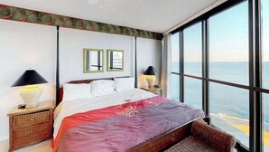 Un dormitorio con una cama grande, dos lámparas de noche, una mesita de noche, una silla de mimbre y vista al océano a través de amplias ventanas.