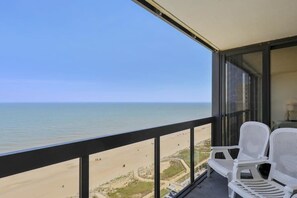 A balcony with a view of the beach and ocean, two white chairs, and a glass door. - Affordable & convenient beachfront condo with balcony, pools & gym (Ocean City)