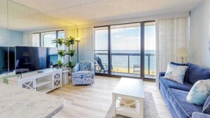 Una sala de estar moderna con televisor, asientos, una mesa de centro y vista a la playa.