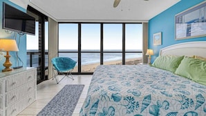 Una camera da letto con un letto, un ventilatore a soffitto, una televisione, una sedia e vista sulla spiaggia.