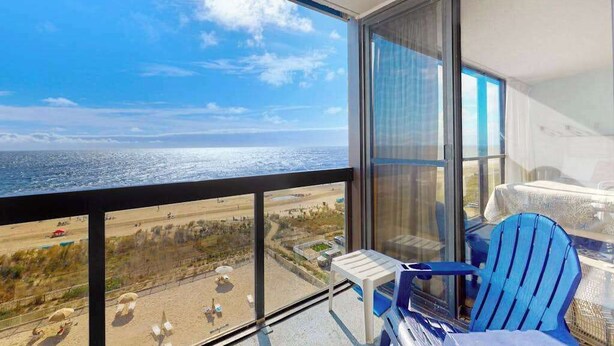 A balcony with a view of the beach and ocean, a table, and two chairs. - Classic, alluring beachfront condo with balcony, views, pools, gym & tennis (Ocean City)