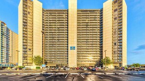 A multi-story hotel with a central vertical feature, a parking lot in front, and a clear blue sky. - Classic, alluring beachfront condo with balcony, views, pools, gym & tennis (Ocean City)