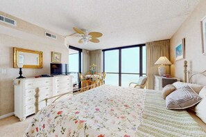 A hotel room with a bed, a desk, a chair, a television, a ceiling fan, and large windows. - Charming condo right on the beach with views, balcony, pools, tennis & gym (Ocean City)