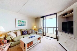 A hotel room with a sofa, a coffee table, a television, and a large window. - Charming condo right on the beach with views, balcony, pools, tennis & gym (Ocean City)