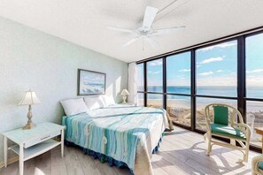 Ein Schlafzimmer mit Bett, Nachttisch, Stuhl, Deckenventilator und großen Fenstern mit Blick auf den Strand.
