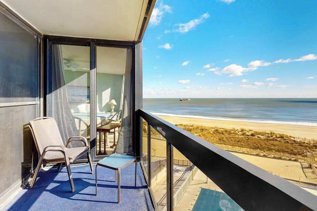 A balcony with a view of the beach, a table, and two chairs.