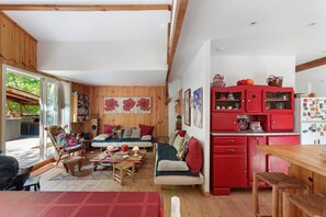 A cozy living room with a sofa, wooden flooring, and a red kitchen area with cabinets. - Holiday Home "Cabane Eden Pyla" with Private Terrace and Garden (La Teste-de-Buch)