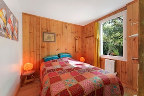 A bedroom with a wooden wall, a bed with a colorful quilt, a small stool, a lamp, and a window with a view of greenery. - Holiday Home "Cabane Eden Pyla" with Private Terrace and Garden (La Teste-de-Buch)