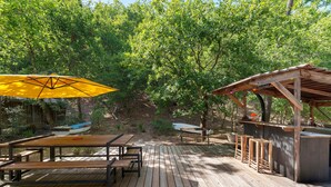 Una terraza de madera al aire libre con mesas de picnic y un paraguas amarillo, rodeada de árboles.