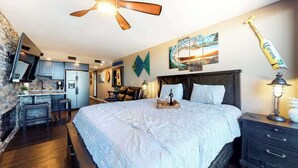 Un dormitorio con cama, escritorio, silla, ventilador de techo, mesita de noche y televisor instalado en la pared.