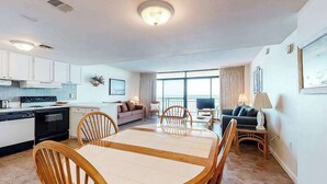 A modern kitchen with white cabinets, a dining area with a round table and chairs, a living room with a sofa and a TV, and large windows offering a view. - Fountainhead Towers 1204 (Ocean City)