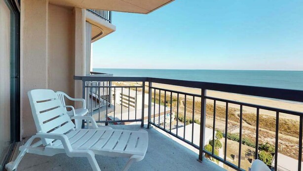 A balcony with two white lounge chairs, a view of the beach, and a clear sky. - Fountainhead Towers 1204 (Ocean City)